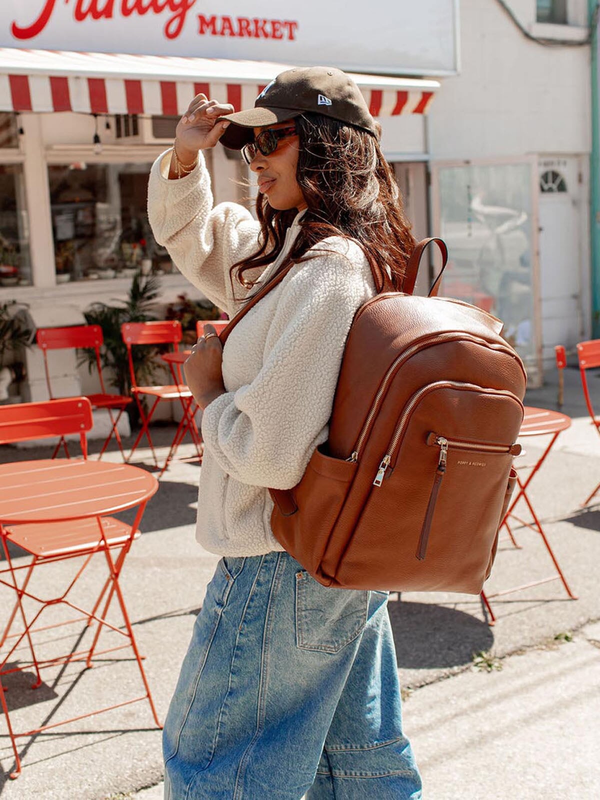 Woman with a brown vegan leather backpack walking past a market with red and white striped awning.