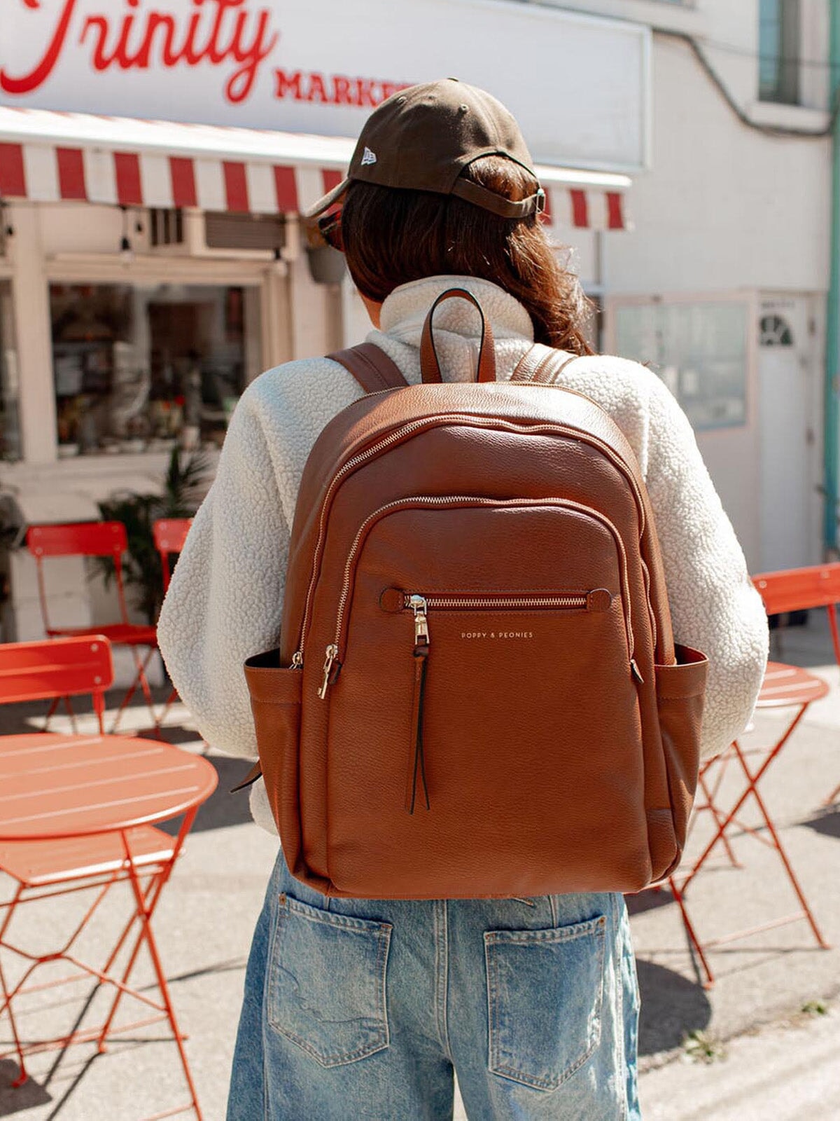 Person wearing a brown vegan leather backpack in an outdoor setting with tables and chairs.
